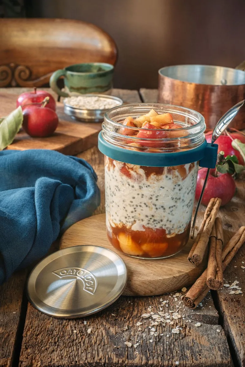 Apple pie overnight oats layered in an OATOLOGY glass jar, showing the cinnamon-stewed apple and creamy oats through the glass