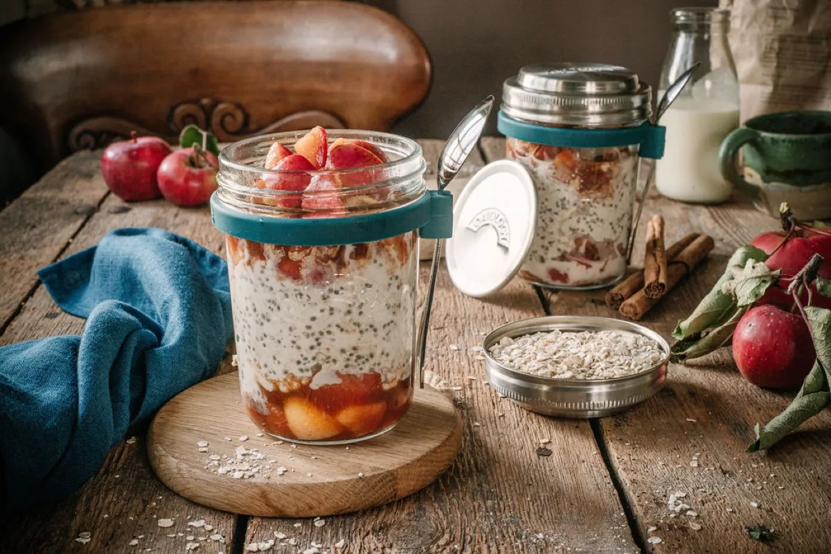 Jar of apple pie overnight oats on a wooden board beside the stainless steel lid and a red apple