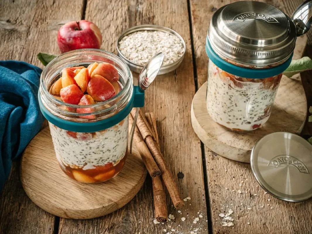 Apple pie overnight oats in an OATOLOGY jar, layered with cinnamon-stewed apple and topped with more stewed apple, next to the stainless steel lid