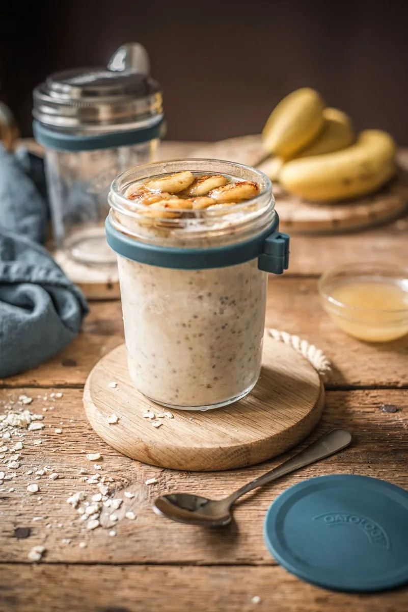 Open jar of banana bread overnight oats topped with golden caramelised banana slices and a dusting of cinnamon, spoon resting inside