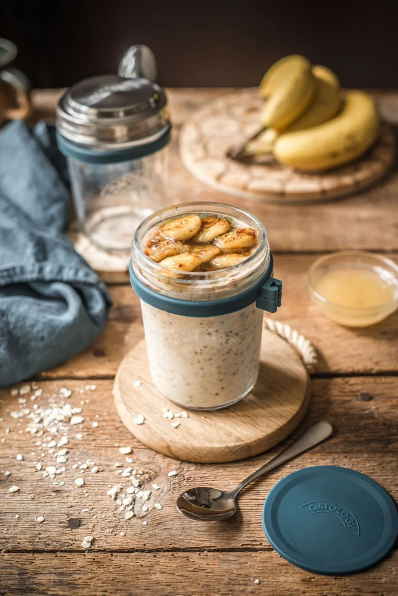 Jar of banana bread overnight oats on a wooden board with the OATOLOGY lid and a ripe spotty banana alongside