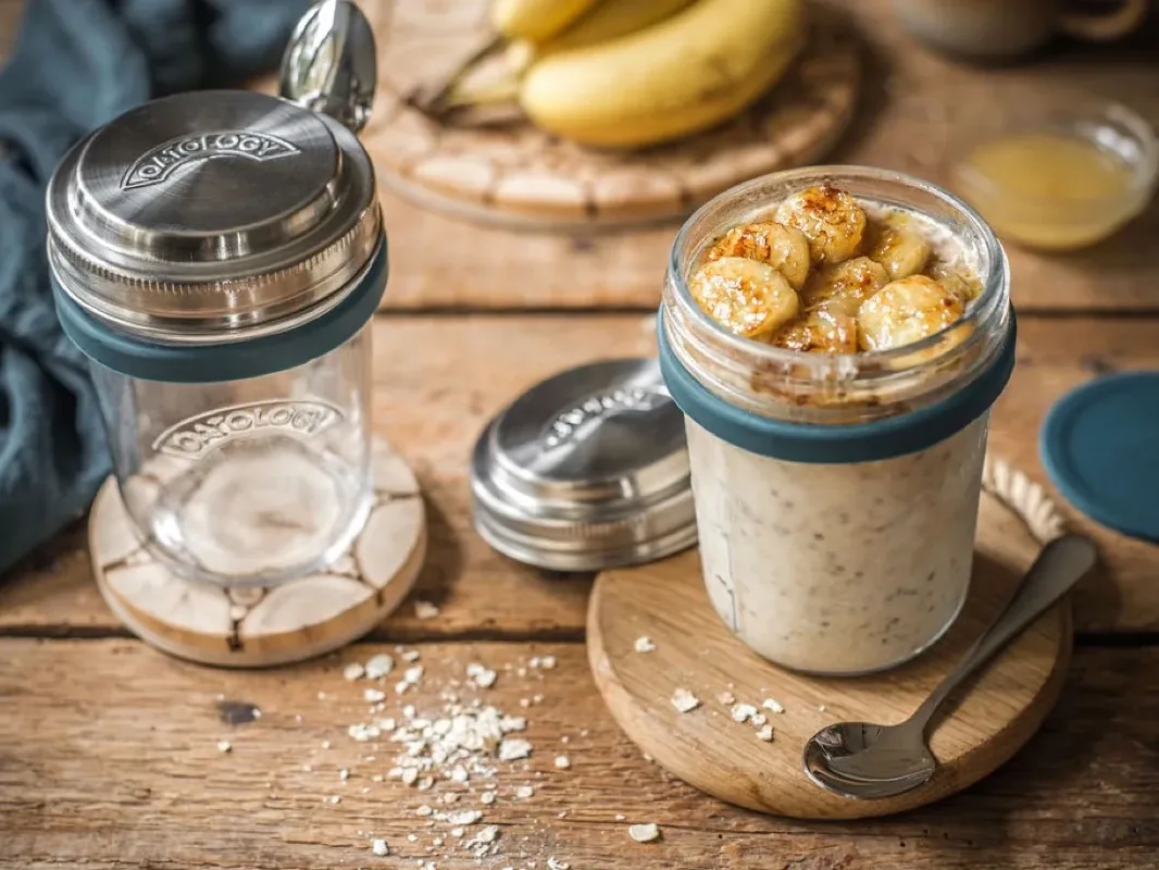 Banana bread overnight oats in an OATOLOGY jar topped with warm caramelised banana slices and a dusting of cinnamon, with a ripe banana behind
