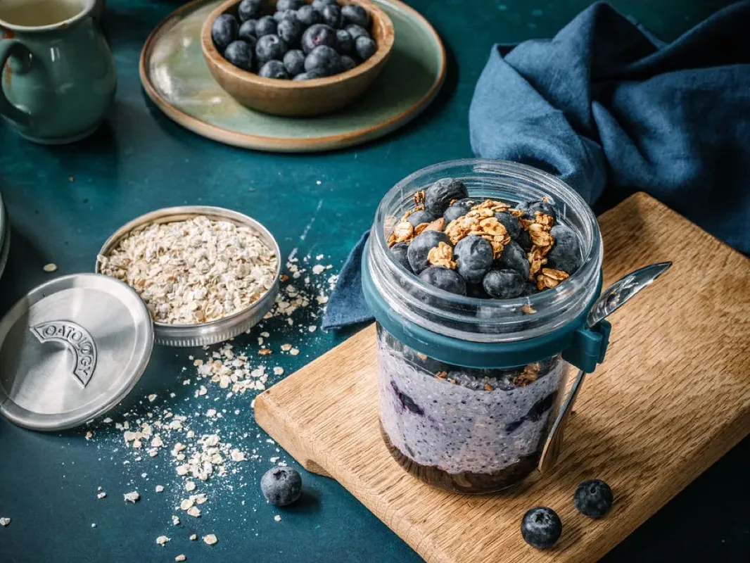 Blended berry overnight oats in an OATOLOGY jar topped with fresh blueberries, next to the stainless steel lid filled with oats