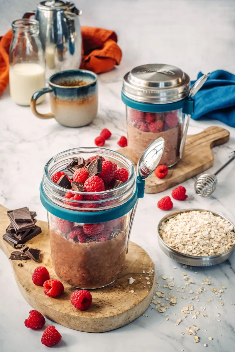 Chocolate and raspberry overnight oats in an OATOLOGY glass jar topped with fresh raspberries and dark chocolate chips, on a wooden board