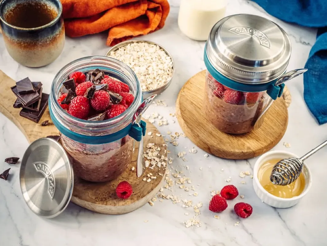 Chocolate and raspberry overnight oats in an OATOLOGY jar topped with fresh raspberries and dark chocolate, next to the stainless steel lid filled with oats