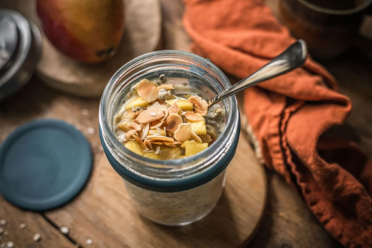 Open jar of mango and coconut overnight oats topped with toasted flaked almonds and diced mango, with a spoon resting inside