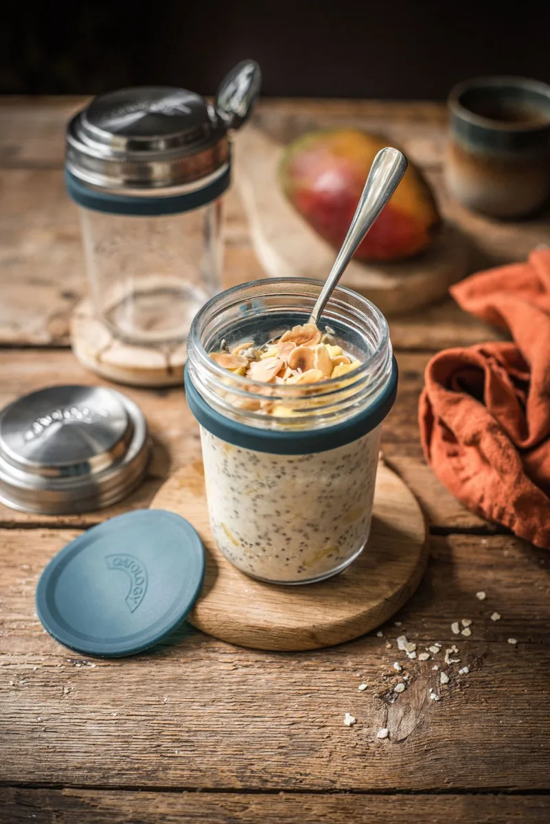 Jar of mango and coconut overnight oats on a wooden board with the OATOLOGY lid, a second empty jar, and a whole mango behind
