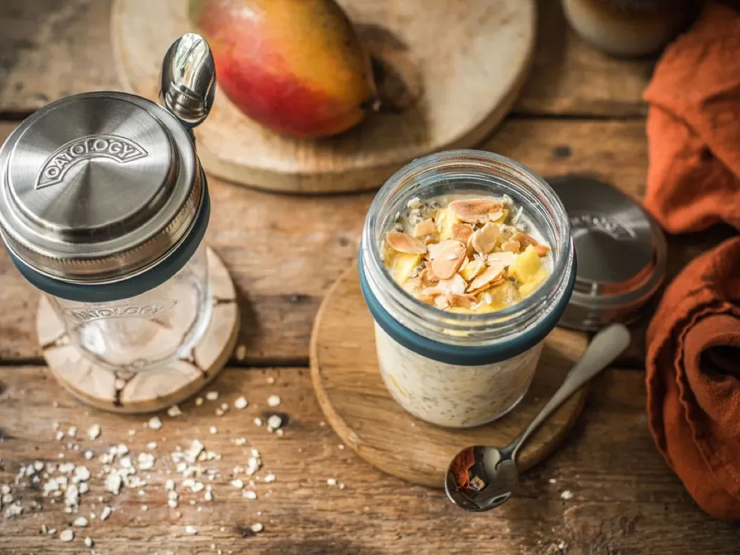Mango and coconut overnight oats in an OATOLOGY jar beside a second jar with the stainless steel lid and clip-on spoon, with a whole mango behind