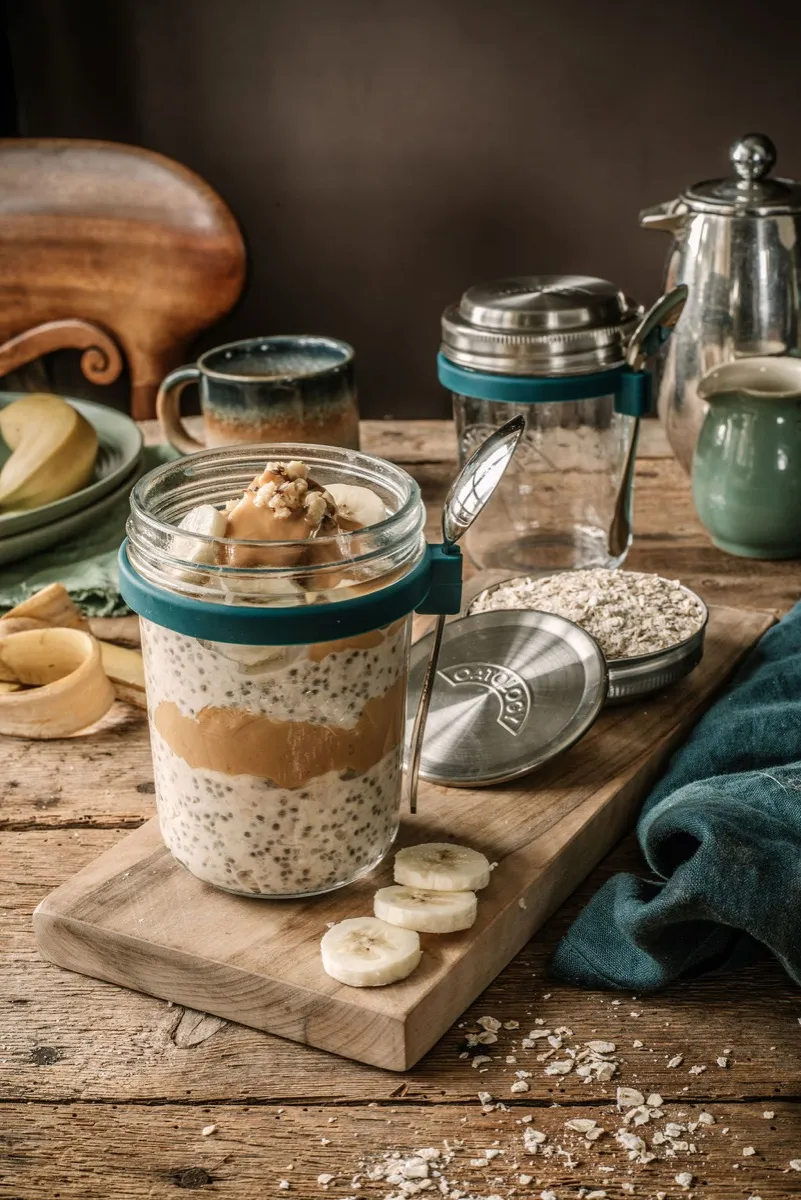 Peanut butter and banana overnight oats layered in an OATOLOGY glass jar, with banana slices on a wooden board