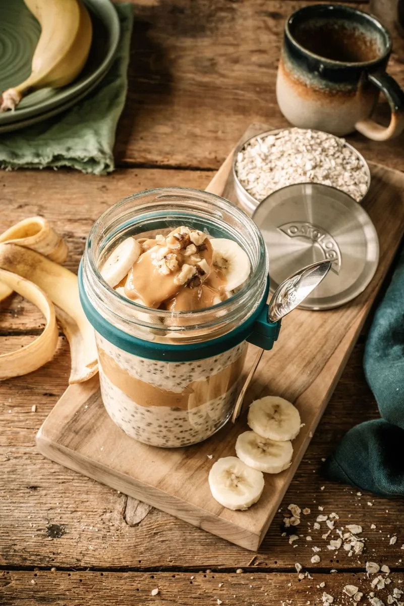 Open jar topped with peanut butter, walnuts, and banana slices, with the spoon clipped to the jar
