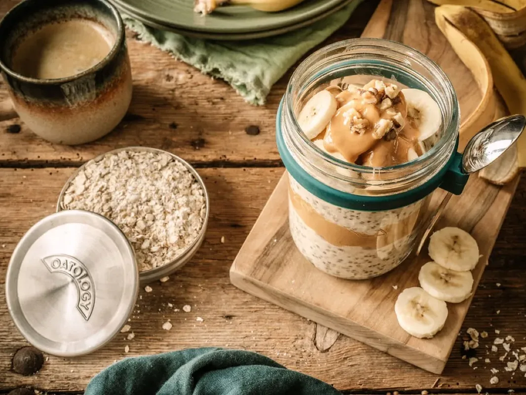 The OATOLOGY stainless steel measuring lid filled with a 50g portion of rolled oats, beside a glass jar of finished overnight oats topped with banana and walnuts