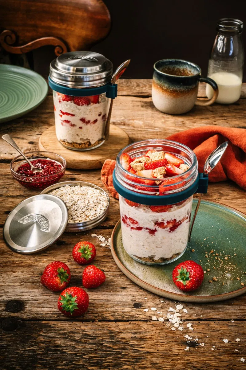 Strawberry cheesecake overnight oats layered in an OATOLOGY glass jar with fresh strawberries on a wooden board